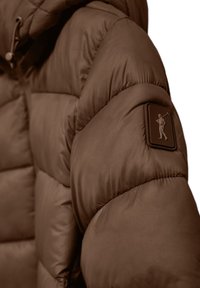 Brun puffjacka med quiltat mönster, med en texturerad axelpatch som avbildar en figur. Slät yta med elasticerade manschetter.