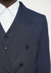 Tengeri kétsoros blazer texturált anyagból, két fekete gombbal és csúcsos hajtókával, fehér csíkos inggel párosítva.