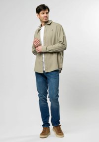 Camisa de botón de manga larga en verde claro sobre una camiseta blanca, combinada con jeans azules y zapatillas marrones con cordones. Diseño sencillo y ajuste casual.