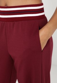 Zbliżenie na osobę noszącą bordowe spodnie z elastycznym, paskowanym pasem, z ręką umieszczoną w prawej kieszeni.