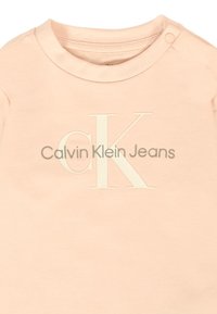 Felpa rosa chiaro con scollatura a girocollo e un bottone a pressione sulla spalla. Logo in rilievo color crema "CK" e "Calvin Klein Jeans" sottostante.