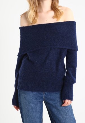 Kvinde iført en marineblå off-shoulder uldtrøje og blå jeans, stående med hænderne afslappet ned langs siden.