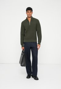Pulover elastan verde închis cu guler cu jumătate de fermoar, purtat cu pantaloni largi bleu marin și pantofi de piele negri, ținând o geantă din piele neagră.