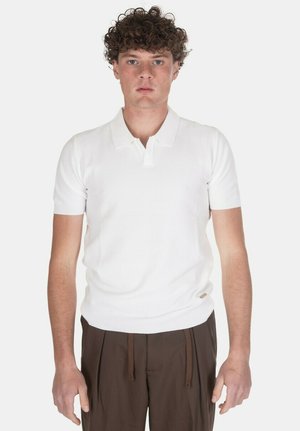 Giovane uomo con capelli ricci che indossa una polo bianca a maniche corte e pantaloni marroni, che guarda avanti su uno sfondo semplice.