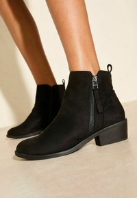 Botas de tobillo de ante negro con cremallera lateral y tacón bajo apilado, llevadas sin calcetines sobre un fondo de suelo y pared clara.