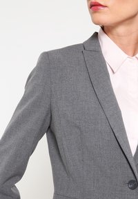 Šedý blazer z texturované látky, se zapínáním na jeden knoflík, klopovým límcem a strukturovanými rameny. Nositý přes světle růžovou košili.