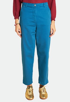 Blaue Baumwollhosen mit gerade geschnittenen Beinen, ausgestattet mit einem vorderen Knopfverschluss und seitlichen Taschen. Kombiniert mit reflektierenden goldenen Sneakers.