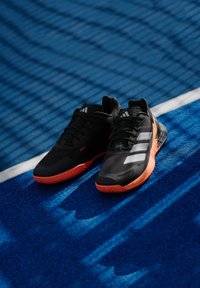 Μαύρα αθλητικά παπούτσια με υφή στο επάνω μέρος, που διαθέτουν τρεις λευκές ρίγες και πορτοκαλί σόλα, τοποθετημένα σε μπλε επιφάνεια γηπέδου.