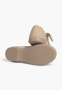Beige Slipschuhe mit einer strukturierten Oberseite, glatter Gummisohle und einem kleinen elastischen Riemen-Detail an der Ferse. Die Sohle weist Griffmuster auf.