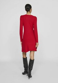 Garais piedurkņu sarkanais ribotais kleitas siluets ir piegulošs, tai ir plecu pogu akcenti un nedaudz paplašināts apakšmalas dizains. Valkāta ar melniem zābakiem līdz ceļgaliem.