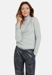 Kobieta z długimi brązowymi włosami, ubrana w jasnoszary sweter z golfem i czarne skórzane spodnie, stojąca z jedną ręką w kieszeni.