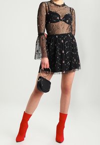 Schwarzes florales Kleid mit transparentem langärmligem Überzug, hohem Kragen, Rüschenmanschetten, kombiniert mit roten Stiefeletten und einer schwarzen Mini-Tasche.