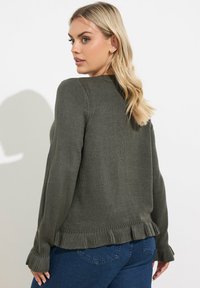 Oliwkowy sweter robiony na drutach z okrągłym dekoltem, ściągaczami na rękawach i falbaną na dolnej krawędzi. Noszony z niebieskimi dżinsami, które prezentują luźny krój.