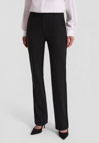 Czarne spodnie w prążki o dopasowanym kroju, z gładką strukturą i elastycznym pasem. Noszone z jasną bluzką i spiczastymi obcasami.