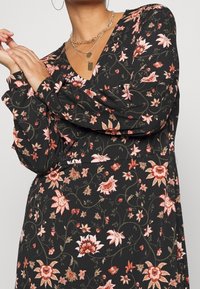 Musta kukkamekko, jossa on pitkät hihat, V-pääntie ja monimutkaisia kukkakuvioita vaaleanpunaisen, punaisen ja vihreän sävyissä. Pehmeä kudottu kangas.