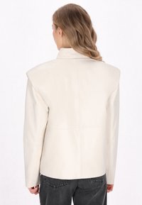 Creme læderjakke med strukturerede skuldre, høj krave og en glat tekstur. Designet har minimal syning og en skræddersyet pasform.