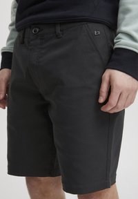 Svart shorts laget av solid stoff; har knappelukking, forklepse lommer og en liten logodetalj på siden.