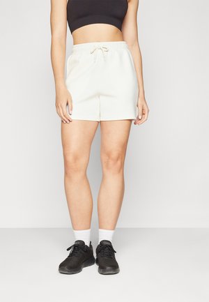 Kremfargede sweatshorts med elastisk midjebånd og snor, med en jevn tekstur og avslappet passform. Svarte joggesko med hvite sokker.