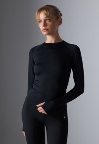 Crna dugorukava sportska majica s pripijenim dizajnom, izrađena od glatkog i rastezljivog materijala. Ima raglan rukave i rupice za palčeve na manžetama.
