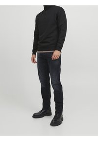 Uomo che indossa un pullover nero con zip a quarto, jeans scuri e stivali in pelle neri con lacci, in piedi contro uno sfondo chiaro e semplice.