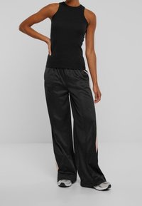 Černý top bez rukávů kombinovaný s volnými černými kalhotami s růžovými bočními pruhy, vyrobený z hladké látky, vhodný pro sportovní oblečení.