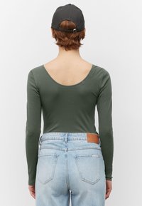 Langärmliger olivgrüner Bodysuit mit einem tiefen Rückendekolleté, kombiniert mit hellblauen High-Waist-Jeans, die mit einem braunen Leder-Logopatch versehen sind.