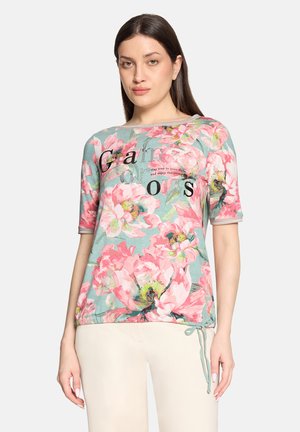 Kvinde iført en turkis blomstret top med pink blomster, sort tekst og beige bukser, stående mod en ensfarvet baggrund.