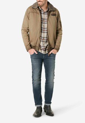 Braune Bomberjacke mit Fellkragen, kariertes Hemd, blaue Jeans und schwarze Stiefel. Verfügt über ein Logo auf der Vorderseite und seitliche Taschen.