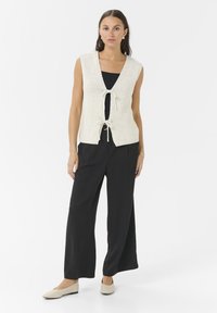 Knit cream vest with a deep V-neck and decorative ties at the waist, paired with black wide-leg trousers and light-colored flats.