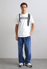 Balta kokvilnas T-kreklu ar melnu "MOSCHINO" tekstu un pīkstošā punktu dizainu, savienots ar brīviem zilajiem džinsiem un melnbaltiem sporta apaviem.