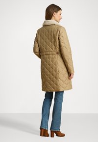 Kvalitatīvs brūns plīvurs ar mākslīgo kažokādu apkakli, vidēja garuma, ar izteiktu jostasvietu, garām piedurknēm un redzamām šuvju detaļām.