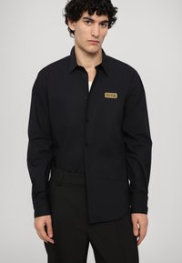 Black long-sleeve button-up shirt made of smooth fabric, featuring a left chest logo patch in gold. Tailored fit with a classic collar.