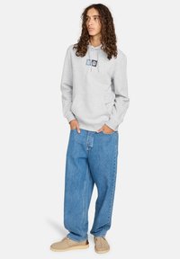 Felpa grigia con tasca frontale e dettaglio grafico nero, abbinata a jeans blu larghi e scarpe beige. Design semplice e vestibilità casual.