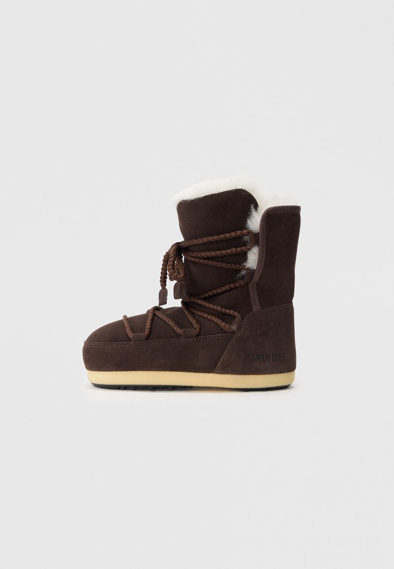 Brun mockaskinnsboot med en vit fuskpälsöverdel, snörning och en tan-färgad gummisula. På sidan står texten "MOON BOOT."