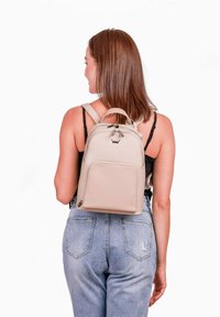 Béžový kožený batoh s kulatým tvarem, dvěma zipy, přední kapsou a nastavitelnými popruhy. Hladká textura, minimalistický design.