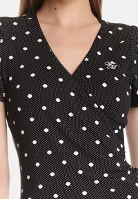 Svart omlottklänning med vita prickar som har cirkulära mönster, V-ringning, korta ärmar och en delikat vågig kant med en liten logotypdetalj.