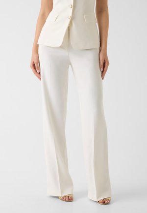 Frau trägt weiße, maßgeschneiderte Hosen mit weitem Bein, kombiniert mit einem passenden ärmellosen Blazer und offenen Sandalen vor einem schlichten Hintergrund.