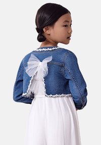 Giacca di denim a vita corta con finiture in pizzo e un fiocco di tulle bianco sul retro, indossata sopra un vestito bianco che rivela una texture stratificata.