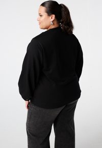 Sweat-shirt noir à coupe décontractée, manches longues, poignets côtelés et col rond, associé à un pantalon large gris foncé.