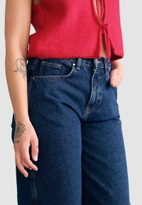 Jeansy dżinsowe z wysokim stanem w ciemnoniebieskim kolorze, z prostymi nogawkami. Cechy to widoczne przeszycia i boczne kieszenie. Akcenty stanowi czerwony dzianinowy top.