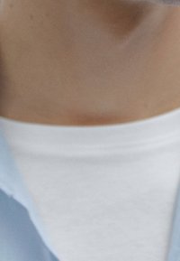 T-shirt bianco sotto una camicia a bottoni blu chiaro. Entrambi i capi sono lisci, con colletto e si adattano bene attorno al collo. Texture sottile visibile.