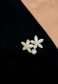 Gold floral brooch featuring two flowers with white petals, one adorned with rhinestones and a central pearl. Background is black fabric.