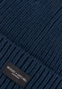 Marineblauwe gebreide muts met geribbelde textuur, met een zwart label waarop "Bickley + Mitchell Amsterdam" op de omslag staat.