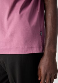 Bras droit d'une personne à la peau foncée portant une chemise rose avec une petite étiquette noire BOSS et un pantalon noir.