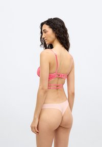 Γυναίκα με σγουρά μαλλιά, φοράει ένα ροζ δαντελένιο σουτιέν με διακοσμητικές τιράντες και ανοιχτό ροζ στρινγκ, στέκεται πλάγια μπροστά σε ένα απλό φόντο.