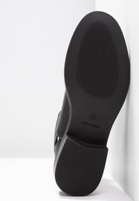 Semelle extérieure de chaussure noire avec une surface en caoutchouc texturée, présentant des crêtes pour la traction et des marquages de la marque. Forme lisse et arrondie.