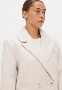 Ανοιχτό μπεζ διπλόσκέπαστο παλτό με υφή μαλακού υλικού, φαρδιές πέτες και μεγάλους κρεμ κουμπιά. Διαθέτει τσέπη στο στήθος στην αριστερή πλευρά.