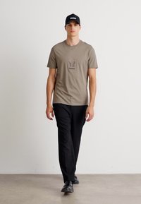 Beige t-skjorte med "HUGO" logo, svarte bukser og en svart caps. Modellen går på en nøytral bakgrunn og viser frem et avslappet antrekk.