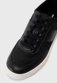 Baskets noires avec un mélange de cuir mat et de tissu texturé. Elles présentent des lacets ronds, des accents cousus et une semelle en caoutchouc blanche.