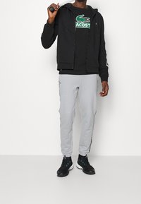 Svart zip-up hoodie och svart t-shirt med grönt Lacoste-logga, matchat med grå mjukisbyxor med sidostråk och svarta sportskor.